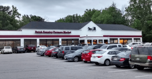 Exterior of Dutch Country Farmers Market with a parking lot filled with cars and trees in the background.