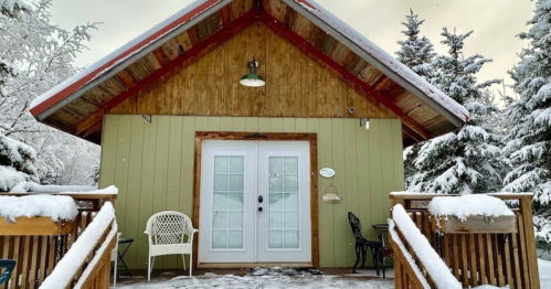 A cozy cabin with a wooden deck, surrounded by snow-covered trees, under a cloudy sky.
