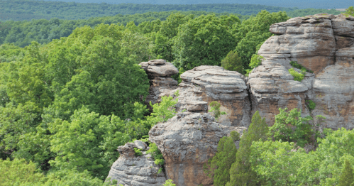 Rock formations surrounded by lush green trees and hills under a clear sky.