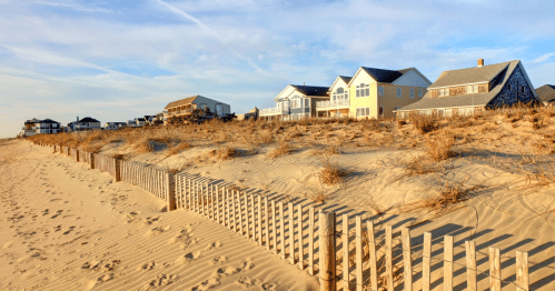 A sandy beach with wooden fencing and houses in the background under a clear blue sky.