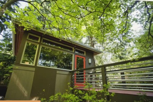 A modern cabin nestled among lush green trees, featuring large windows and a striking red door.