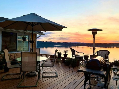 A serene lakeside deck at sunset, featuring a table, chairs, and a patio heater, with calm water reflecting the colors.