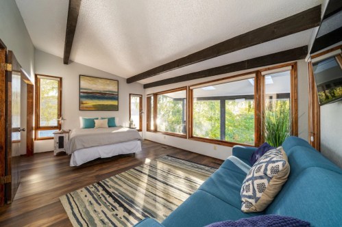 Bright bedroom with large windows, a cozy bed, and a blue sofa, surrounded by greenery and natural light.