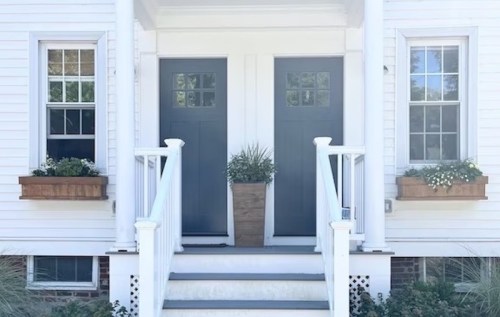 A charming white house with two dark blue doors, flanked by flower boxes and a potted plant on the steps.