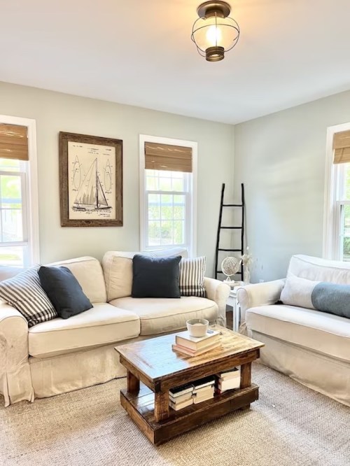 Cozy living room with a light sofa, dark pillows, wooden coffee table, and nautical wall art. Bright natural light.