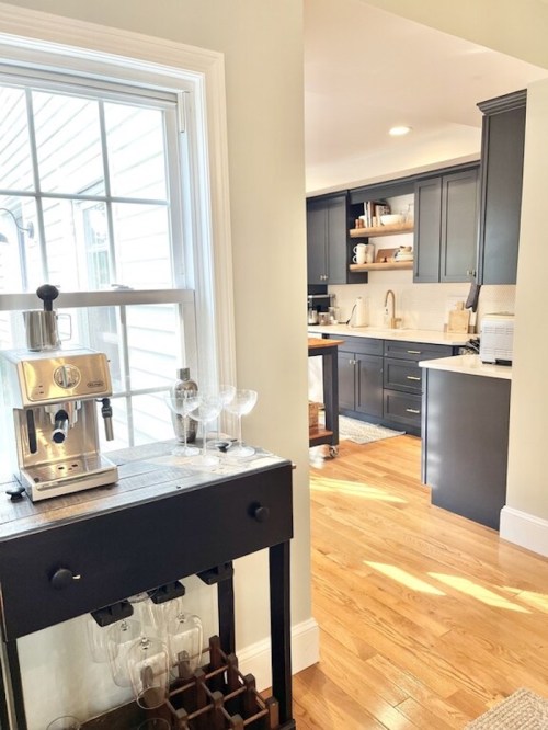 A bright kitchen with dark cabinets, a coffee machine, and a view of a sunny window. Wood floors add warmth.