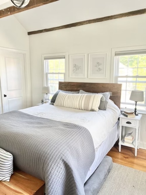Cozy bedroom with a wooden bed, gray bedding, and natural light from windows, featuring a minimalist decor style.