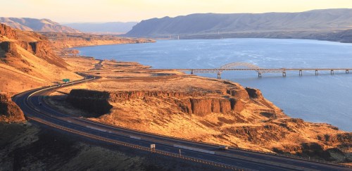 A scenic view of a winding road alongside a river, with cliffs and a bridge in the background during golden hour.