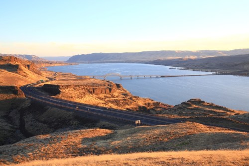 A scenic view of a river winding through hills, with a bridge and road visible under a golden sunset.