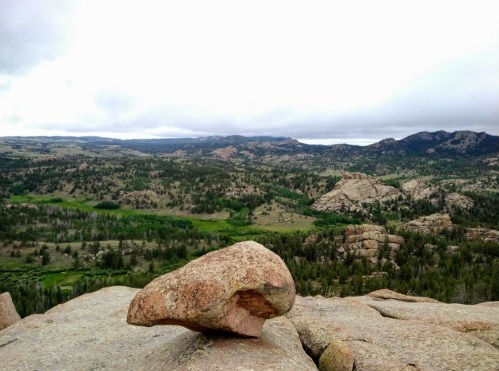A rocky outcrop in the foreground overlooks a lush, green valley and distant mountains under a cloudy sky.