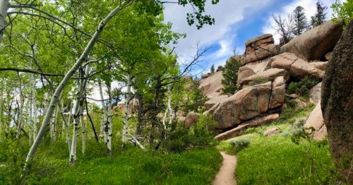 A winding path through lush greenery and aspen trees, leading to rocky outcrops under a partly cloudy sky.