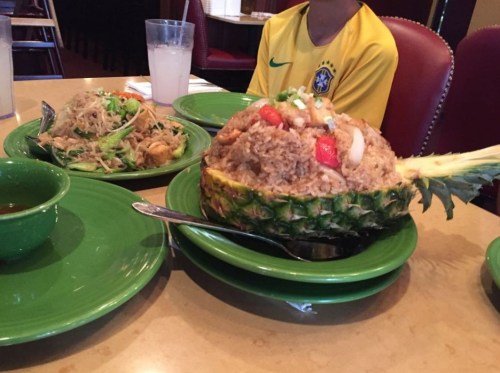 A pineapple filled with fried rice sits on a green plate, alongside a bowl of noodles and a salad.