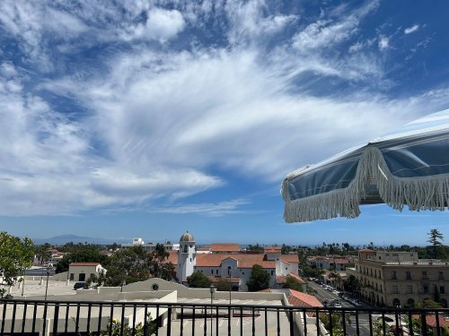 A scenic view of a city skyline under a blue sky with wispy clouds, featuring rooftops and a decorative umbrella.