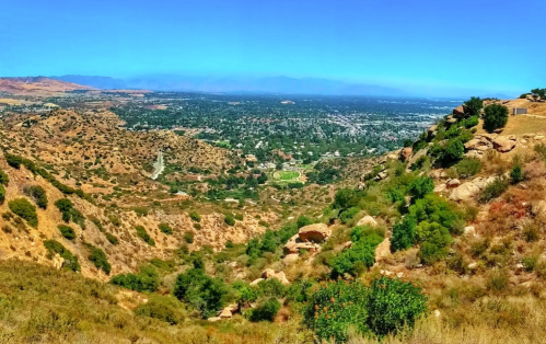 A panoramic view of a valley with hills, greenery, and a distant cityscape under a clear blue sky.