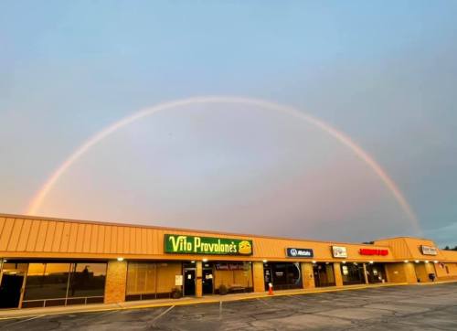 A vibrant rainbow arcs over a shopping plaza with various storefronts under a pastel sky.