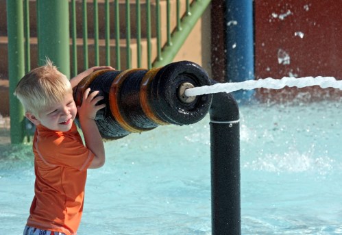 A young boy in an orange shirt joyfully plays with a water spray feature at a pool.