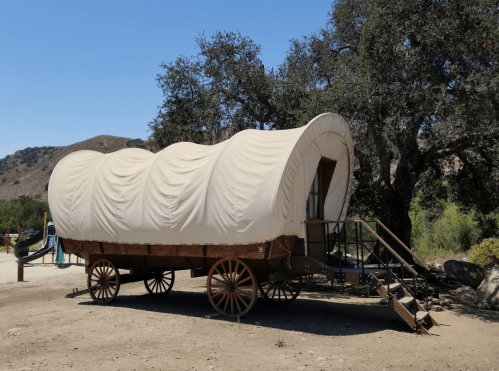 A covered wagon on wheels, set in a sunny outdoor area with trees and a playground in the background.