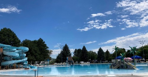 A sunny outdoor pool area featuring water slides, lounge chairs, and trees under a blue sky with scattered clouds.