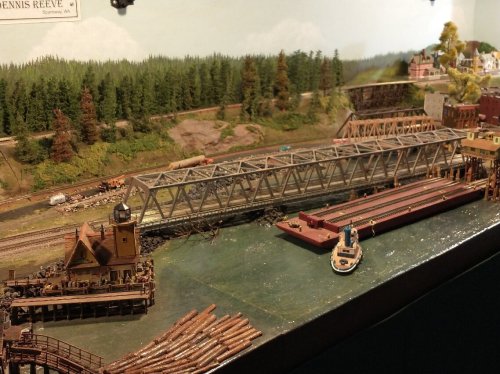 A detailed model train scene featuring a bridge, boats, and a forested backdrop.