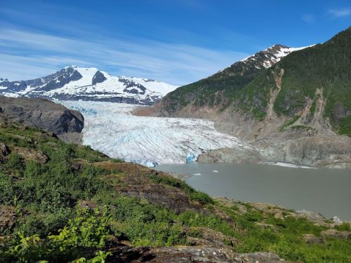 A stunning glacier flows down a mountain, surrounded by rocky terrain and a calm lake under a clear blue sky.