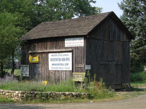 A rustic wooden building with various signs, including one for Weston Little League registration, surrounded by greenery.