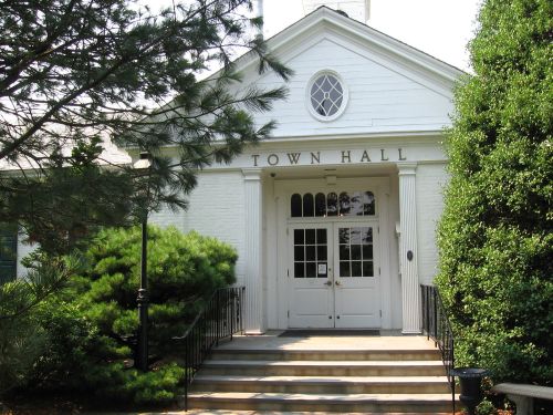 White town hall building with a front entrance, surrounded by greenery and trees.