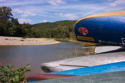 Canoes stacked by a riverbank, surrounded by lush greenery and a clear blue sky.