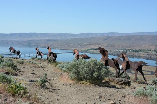 A scenic view of metal horse sculptures on a hillside overlooking a river and mountains in the background.