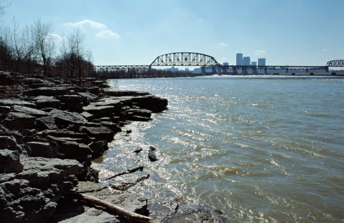A riverbank with rocky shorelines, a bridge in the background, and a city skyline under a clear blue sky.