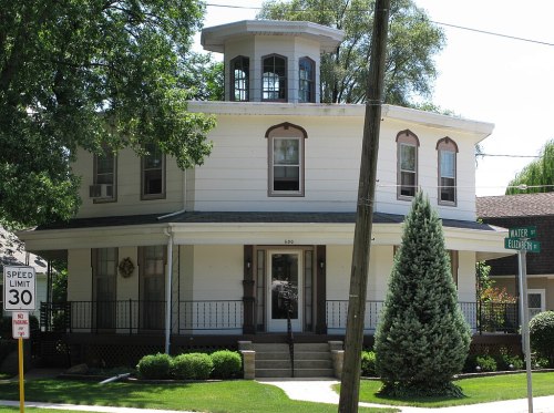 A large, two-story house with a unique octagonal tower and a well-maintained lawn, located at a street corner.