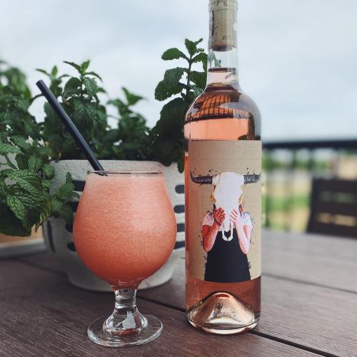 A glass of pink slushy drink next to a bottle of rosé wine featuring a unique label, with a mint plant in the background.