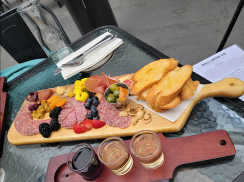 A wooden platter with assorted meats, cheeses, fruits, nuts, and toasted bread, accompanied by small glasses of liquid.