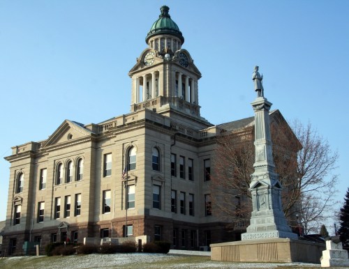 Historic courthouse with a clock tower and a statue in front, set against a clear blue sky. Snow on the ground.