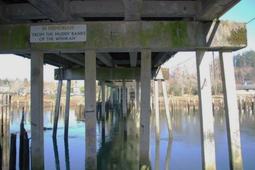 View of a weathered pier with a sign reading "In Memoriam" above the water, surrounded by trees and reflections.