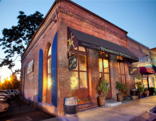 Exterior of a brick building with large windows, featuring a sign for "Woodbridge Uncorked" and potted plants.