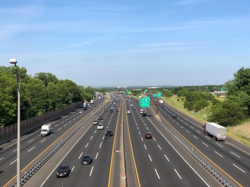 A view of a busy highway with vehicles traveling in both directions, surrounded by greenery and clear blue skies.