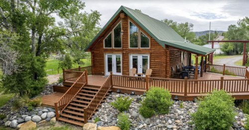 A cozy log cabin with a green metal roof, surrounded by greenery and a wooden deck with seating.