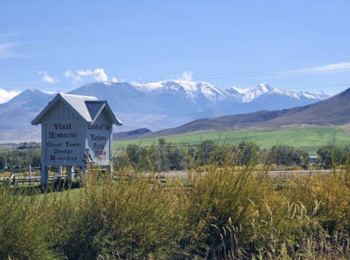 A sign for a historic site with snow-capped mountains in the background and green fields in the foreground.