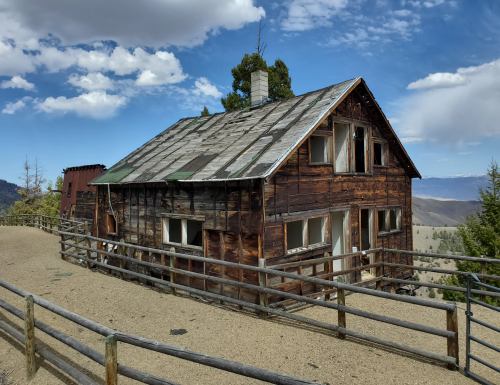 An old, weathered wooden house with a slanted roof, surrounded by a wooden fence and mountains in the background.