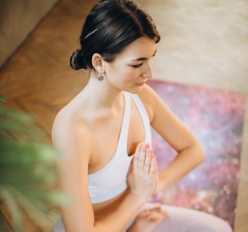 A young woman in a white top sits in a yoga pose, hands in prayer position, with a serene expression.