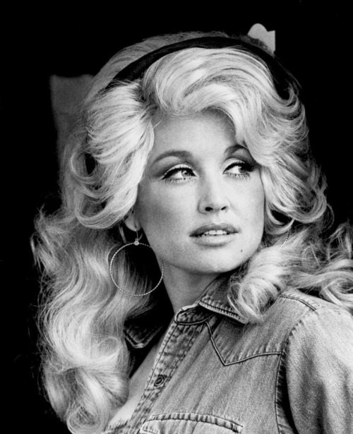 Black and white portrait of a woman with voluminous blonde hair, wearing hoop earrings and a denim shirt, looking thoughtfully.