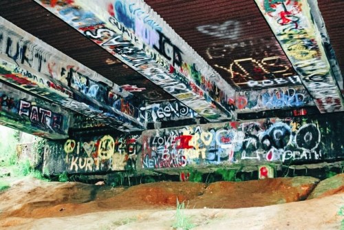 Colorful graffiti covers the underside of a bridge, with various tags and designs on concrete pillars and walls.