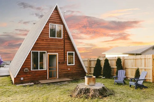 A cozy A-frame cabin with a wooden exterior, surrounded by a fenced yard and chairs, set against a colorful sunset.