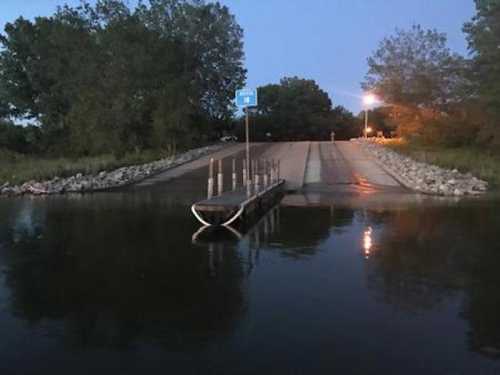 A boat ramp at dusk, with a sign and trees in the background, reflecting in the calm water.