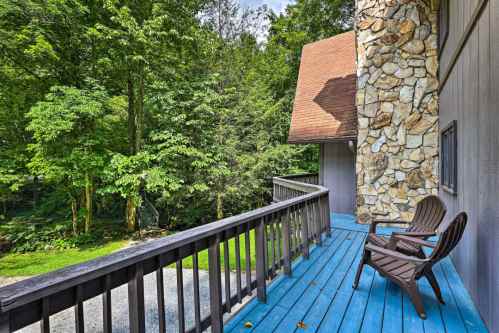 A wooden deck with blue flooring and two chairs, surrounded by lush green trees and a stone wall building.
