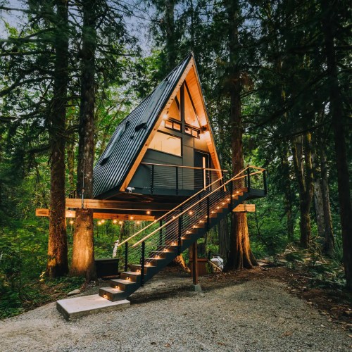 A modern A-frame cabin nestled among tall trees, featuring a staircase and warm outdoor lighting.