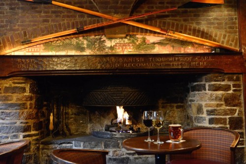 Cozy fireplace in a rustic setting with wine glasses and a beer on a table, surrounded by stone walls and wooden decor.