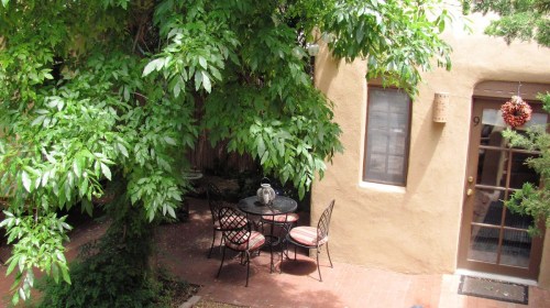 A cozy outdoor patio with a table and chairs surrounded by lush green foliage and a warm, earthy wall.