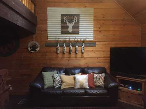 Cozy cabin living room with a brown leather sofa, decorative pillows, wooden walls, and a TV. Antler decor above the sofa.