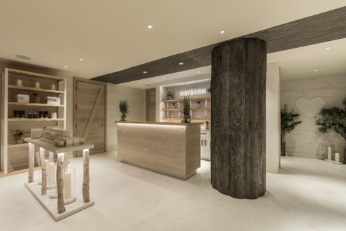 A modern spa reception area with wooden decor, a central column, and shelves displaying candles and wellness products.